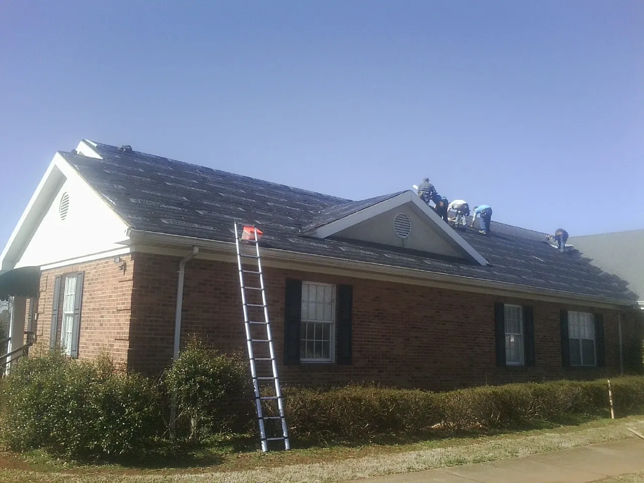 Crew tearing shingles off a brick ranch roof with a ladder set — Metal Roof Repair work by our crew in Smiths Station