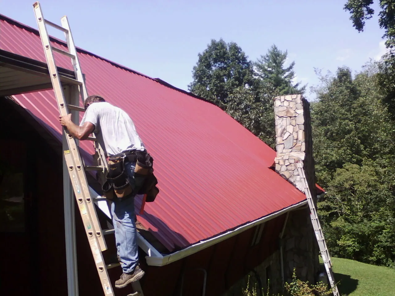 Roofer on a ladder beside a red metal roof and stone chimney — Soffit & Fascia Repair by Roofing Brothers in Smiths Station